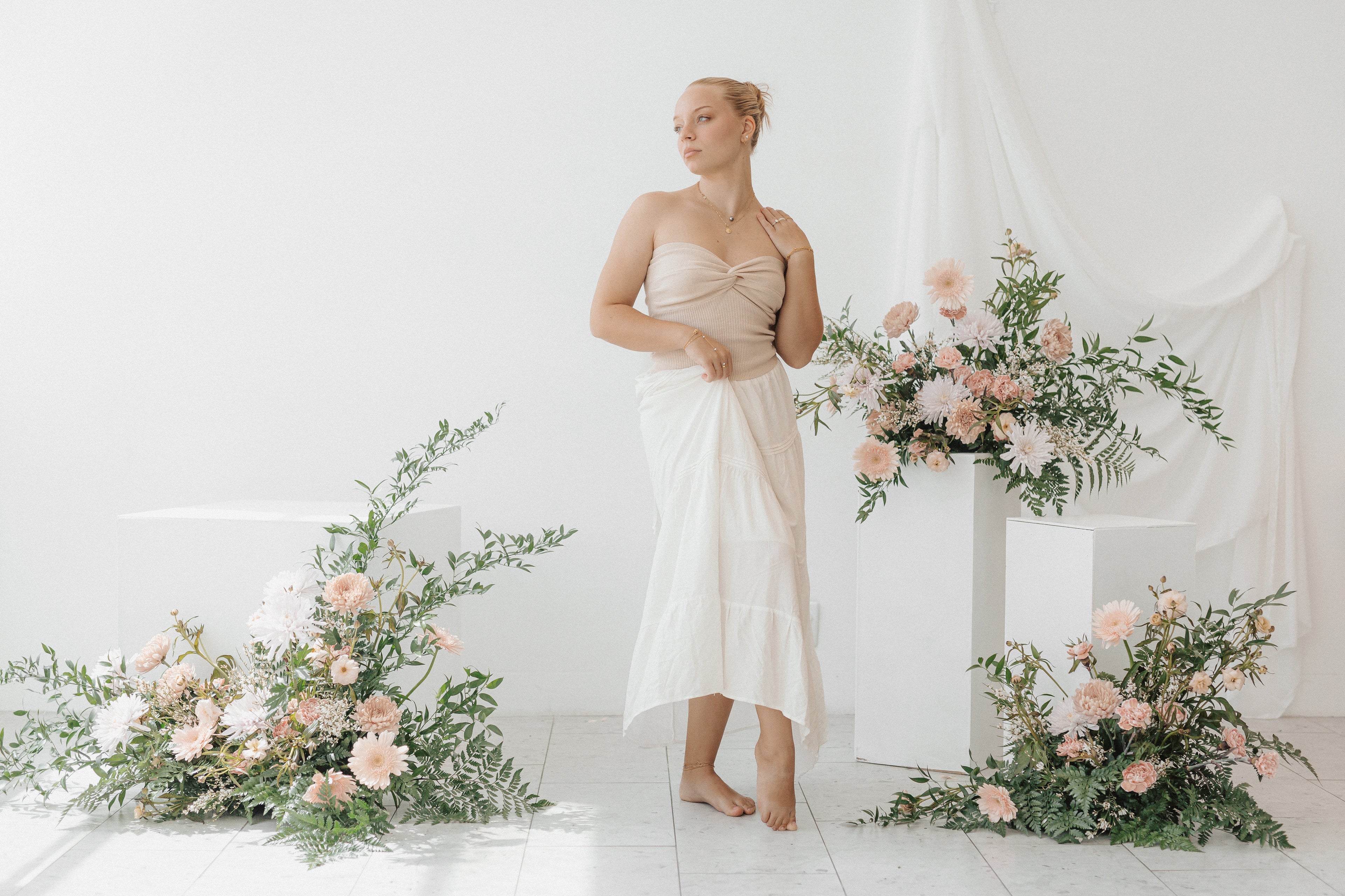 Woman in a beige dress standing between floral arrangements in a white room